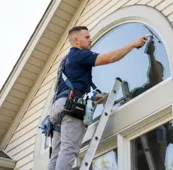 Professional window cleaner on a ladder using a squeegee to clean an exterior arched window, Boca Raton, FL.